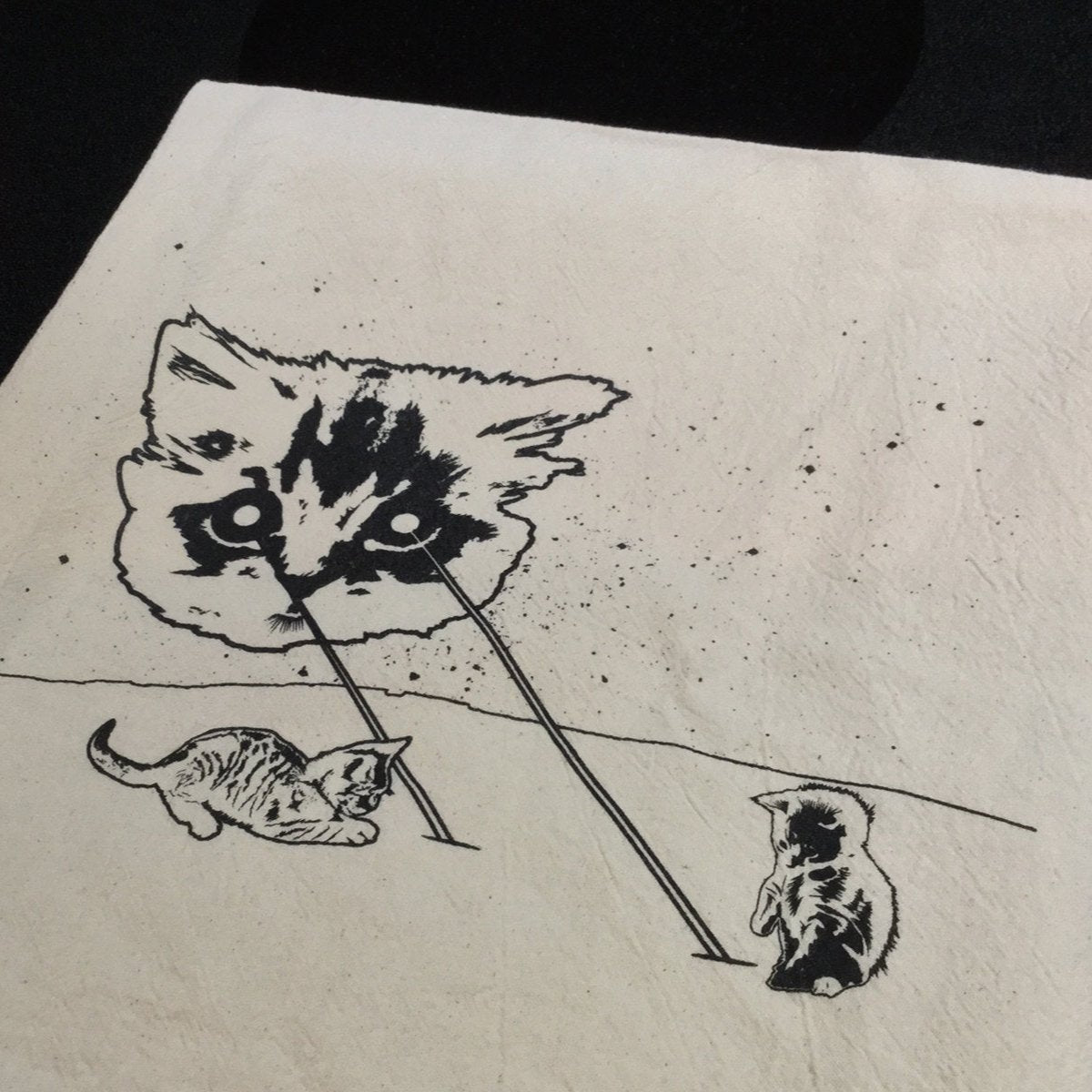 This natural color kitchen towel has a space cat shooting lasers at tiny kitties. The kittens are playing in the lasers. The towel is printed with black ink and is folded on top of a black stove.