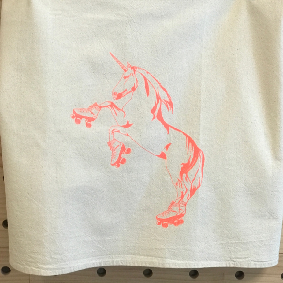 A natural color kitchen towel printed with our roller skating unicorn design in hot pink ink. It is hanging in front of a wooden pegboard wall.