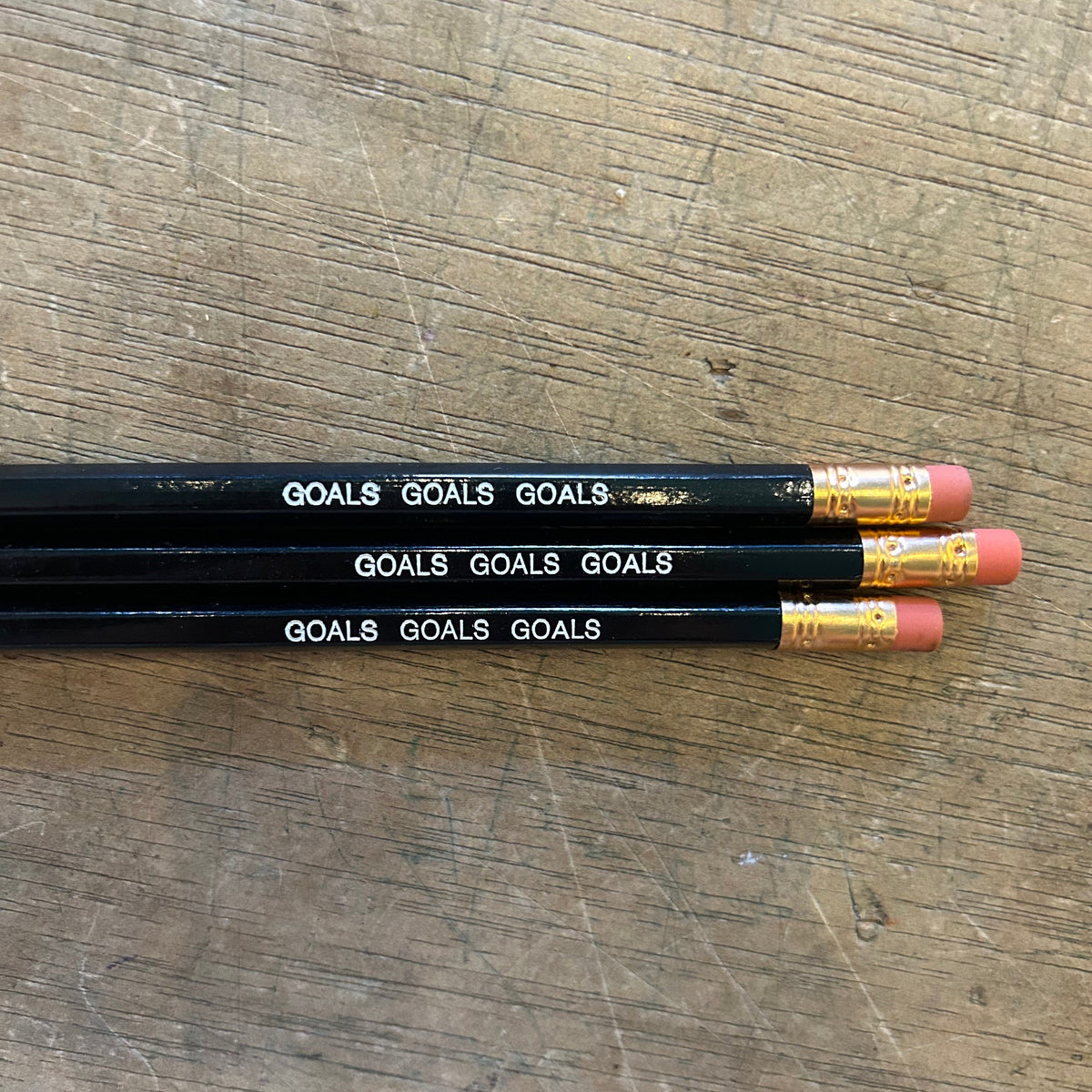 Three black pencils with white 'GOALS' text and gold ferrules, lying on a scratched wooden surface.