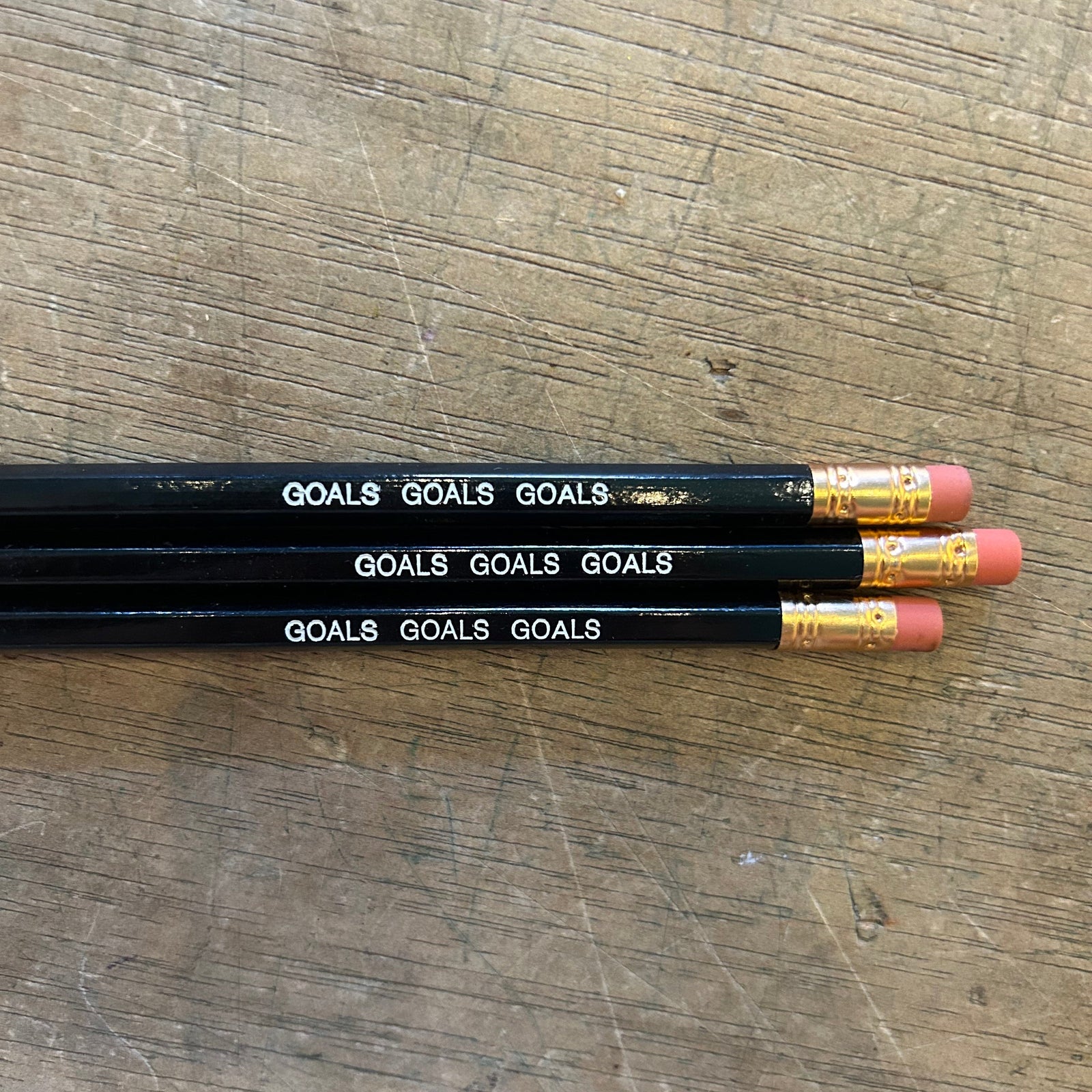 Three black pencils with white 'GOALS' text and gold ferrules, lying on a scratched wooden surface.