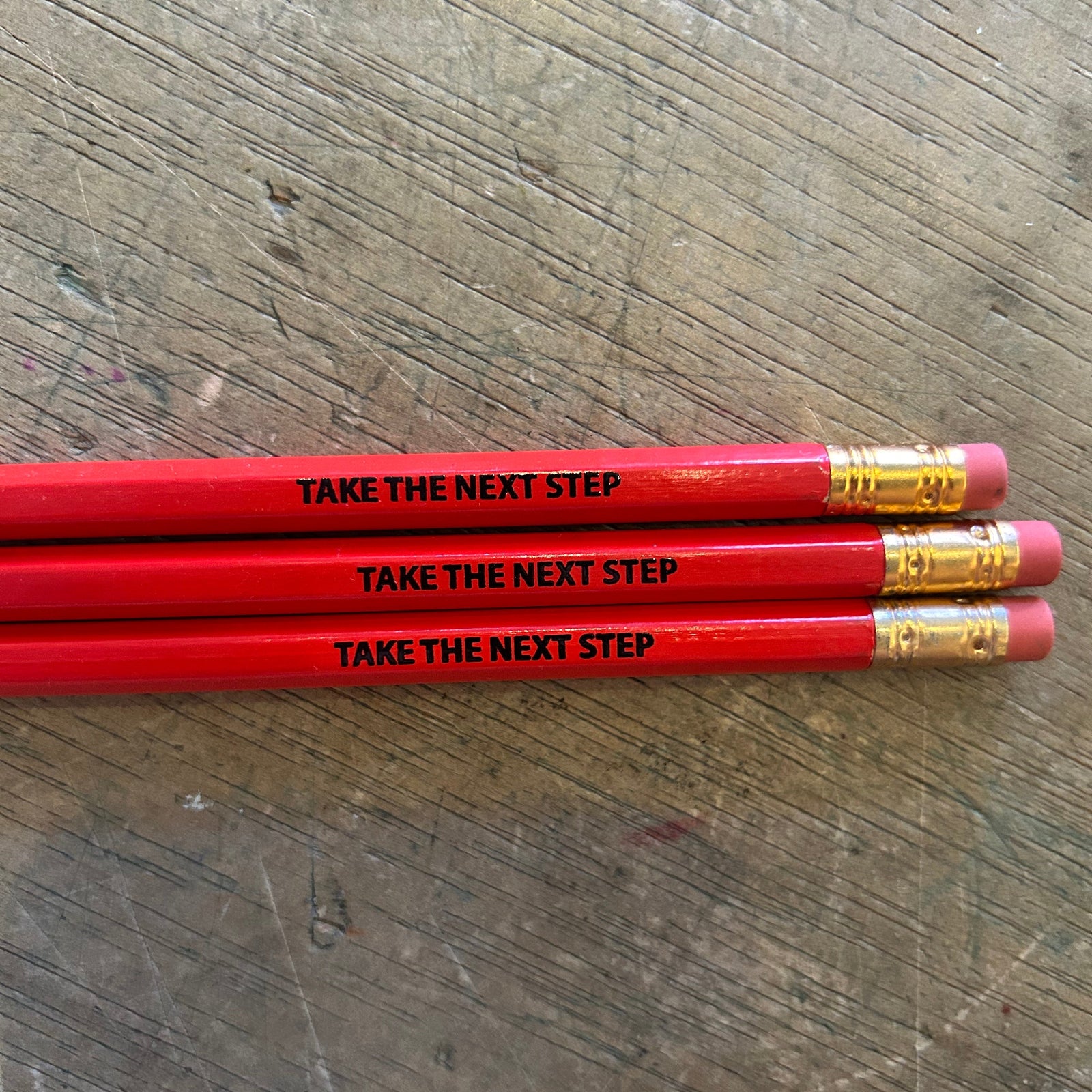 Three red pencils with the phrase 'TAKE THE NEXT STEP' printed in black on each barrel, arranged horizontally on a scratched wooden surface.