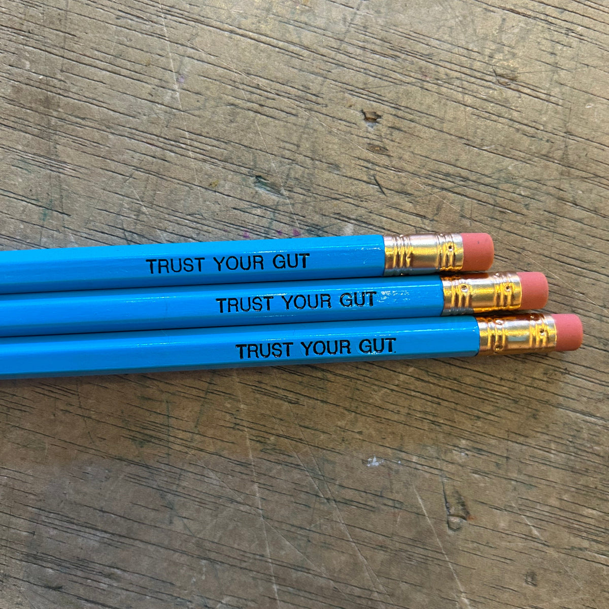 Three blue pencils with black engraved text that says "trust your gut" on a wooden surface. 