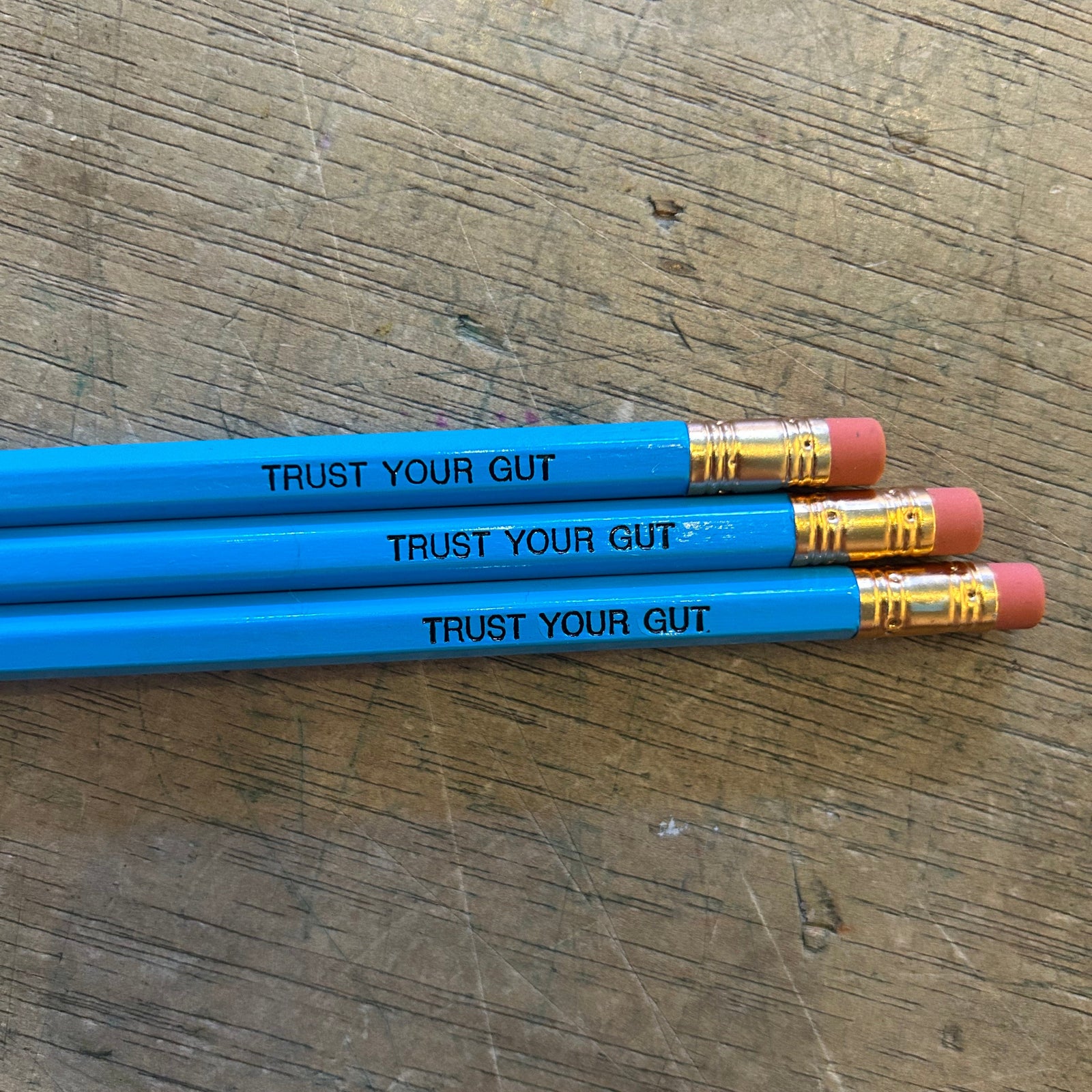 Three blue pencils with black engraved text that says "trust your gut" on a wooden surface. 