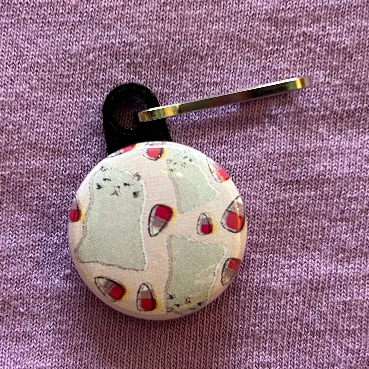 A round, pink zipper pull with a design of three ghost cats and pink candy corn against a purple background. 