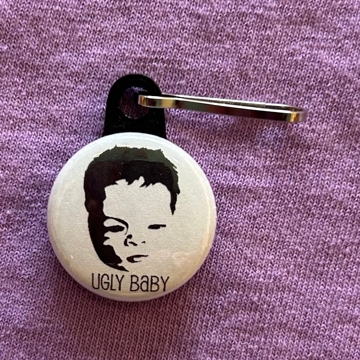 A round, white zipper pull with a baby's face and the text "Ugly Baby" in black on a purple background. 