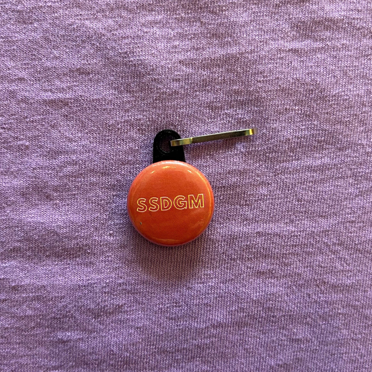 A round, red zipper pull with white outlined text that says "SSDGM" on a purple background. 