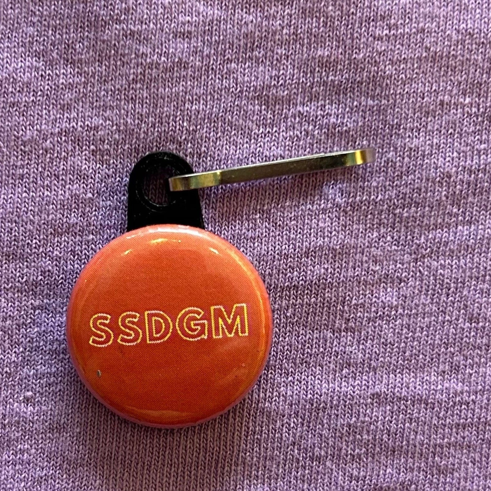 A round, red zipper pull with white outlined text that says "SSDGM" on a purple background. 