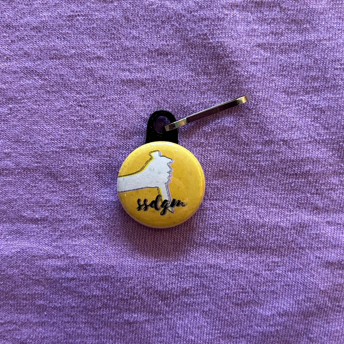 A round, yellow zipper pull with the outline of a hand holding a knife and "ssdgm" in cursive script against a purple background.