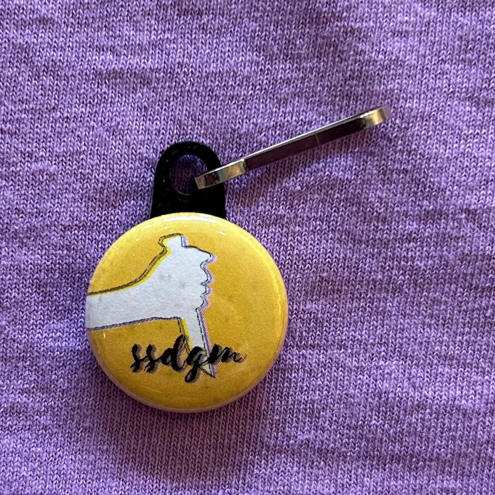 A round, yellow zipper pull with the outline of a hand holding a knife and "ssdgm" in cursive script against a purple background.
