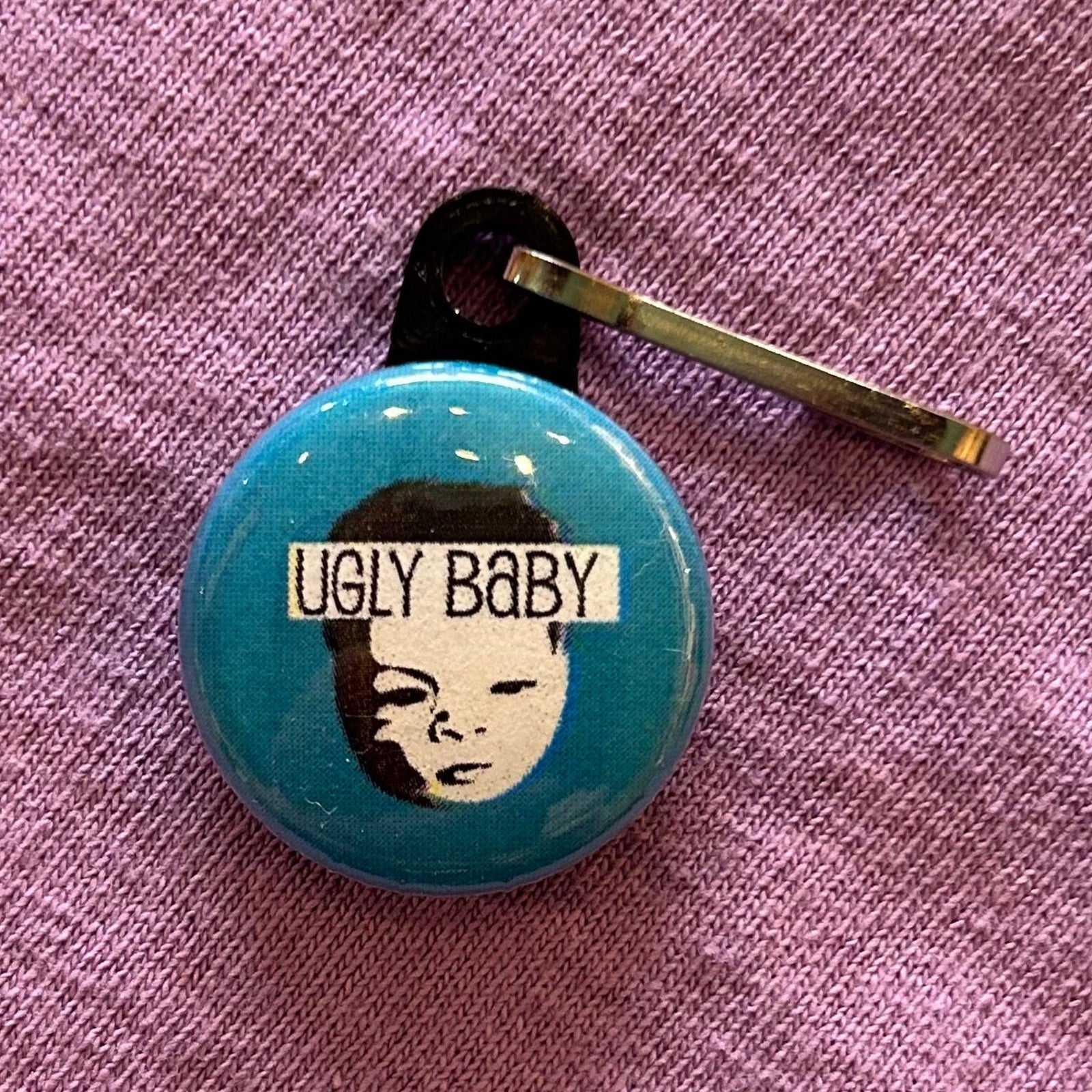 A zipper pull featuring the Ugly Baby logo and text that says "Ugly Baby" on a blue background. 