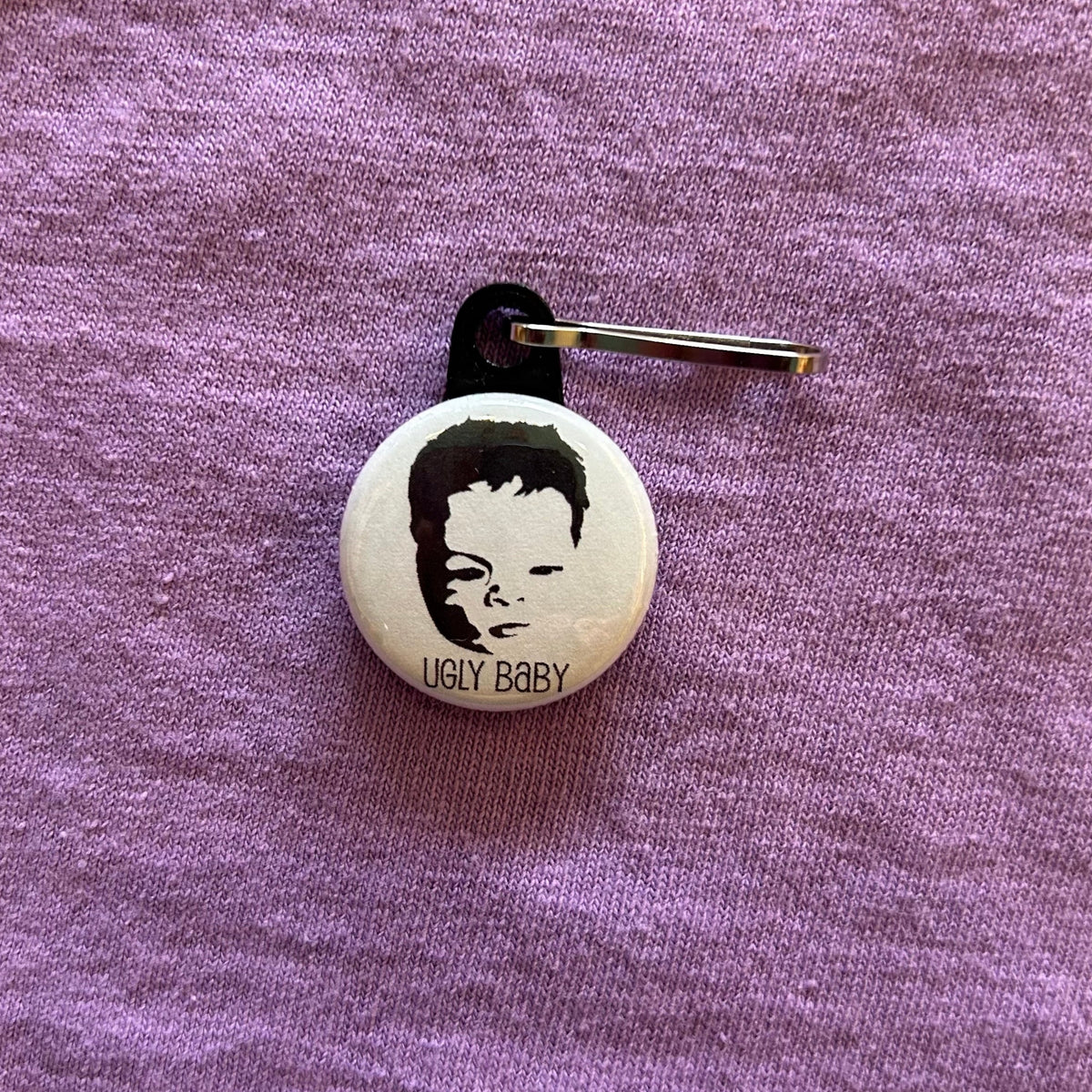 A round, white zipper pull with a baby's face and the text "Ugly Baby" in black on a purple background. 