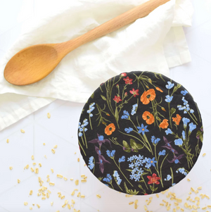 Floral-patterned bowl cover on a white surface with a wooden spoon and scattered seeds.