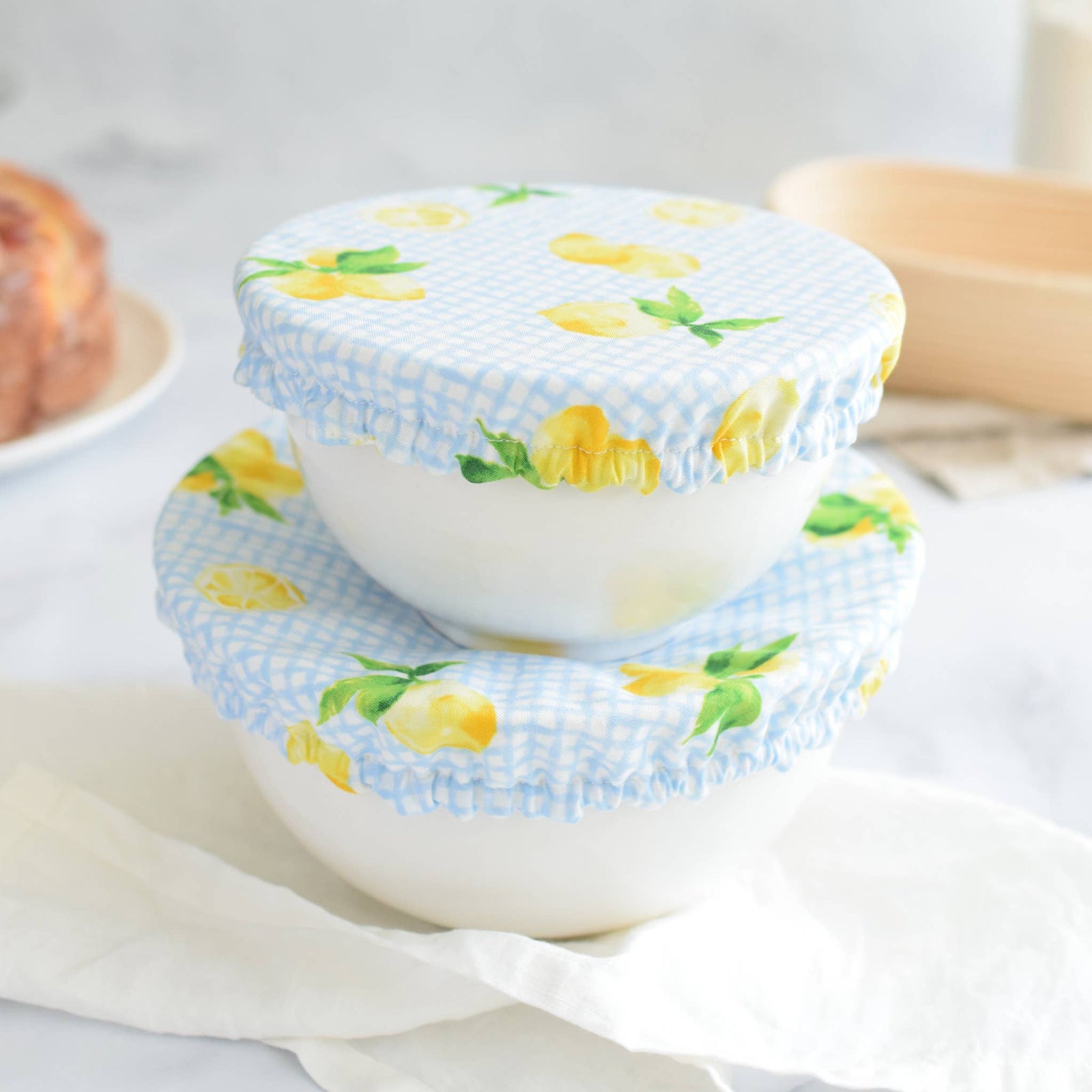 Two white ceramic bowls stacked on top of each other, covered with light blue and white gingham fabric lids printed with yellow lemons and green leaves, set on a white cloth in a softly blurred kitchen setting.