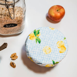 A round reusable fabric storage container lid with a light blue gingham pattern and yellow lemon print, shown on a white surface next to a glass jar of oats, a bowl of oats, walnuts, and an apple.