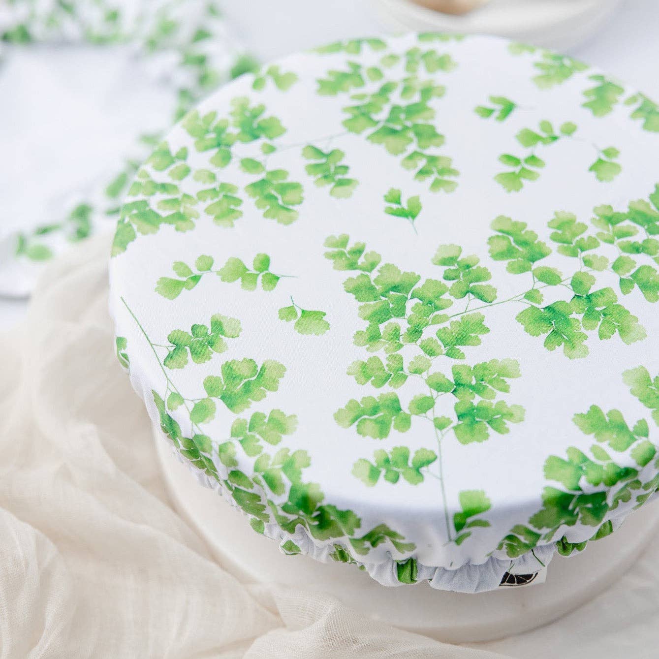 A round white fabric pot cover with a green fern leaf pattern, draped over a white pot on a beige fabric surface.