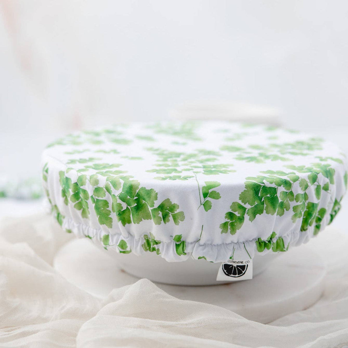 A round white fabric cover with a green leaf pattern, draped over a white bowl on a soft white surface.