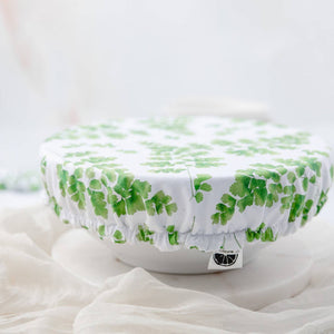A round white fabric cover with a green leaf pattern, draped over a white bowl on a soft white surface.