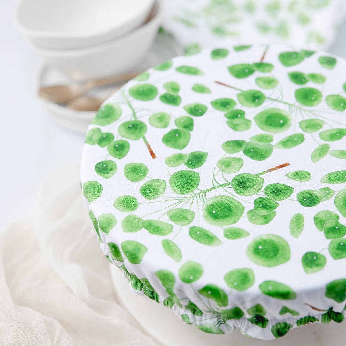 A round fabric storage cover with a green watercolor leaf pattern on a white background, shown draped over a container in a softly styled kitchen setting.