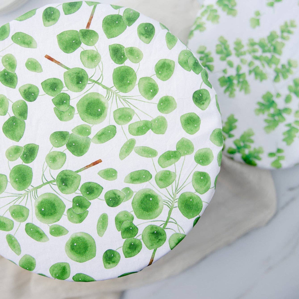 A round white plate or tray with a green watercolor leaf pattern, shown on a light neutral surface with another matching patterned item partially visible in the background.