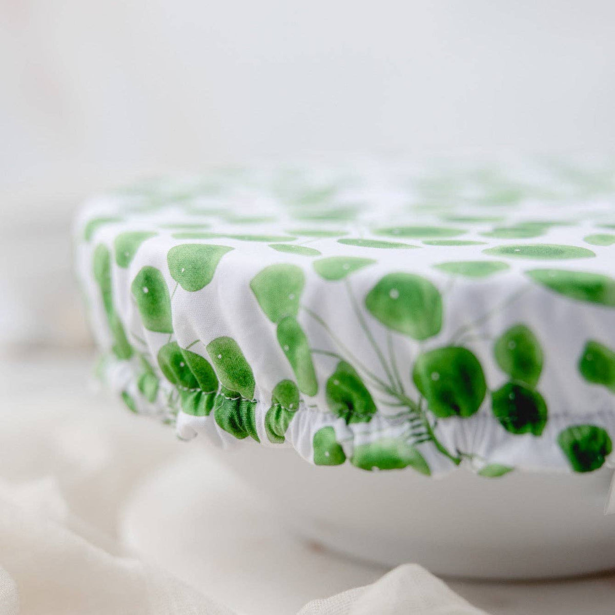 A close-up of a white fabric cover with a green leaf pattern stretched over a round container, set against a soft, light background.
