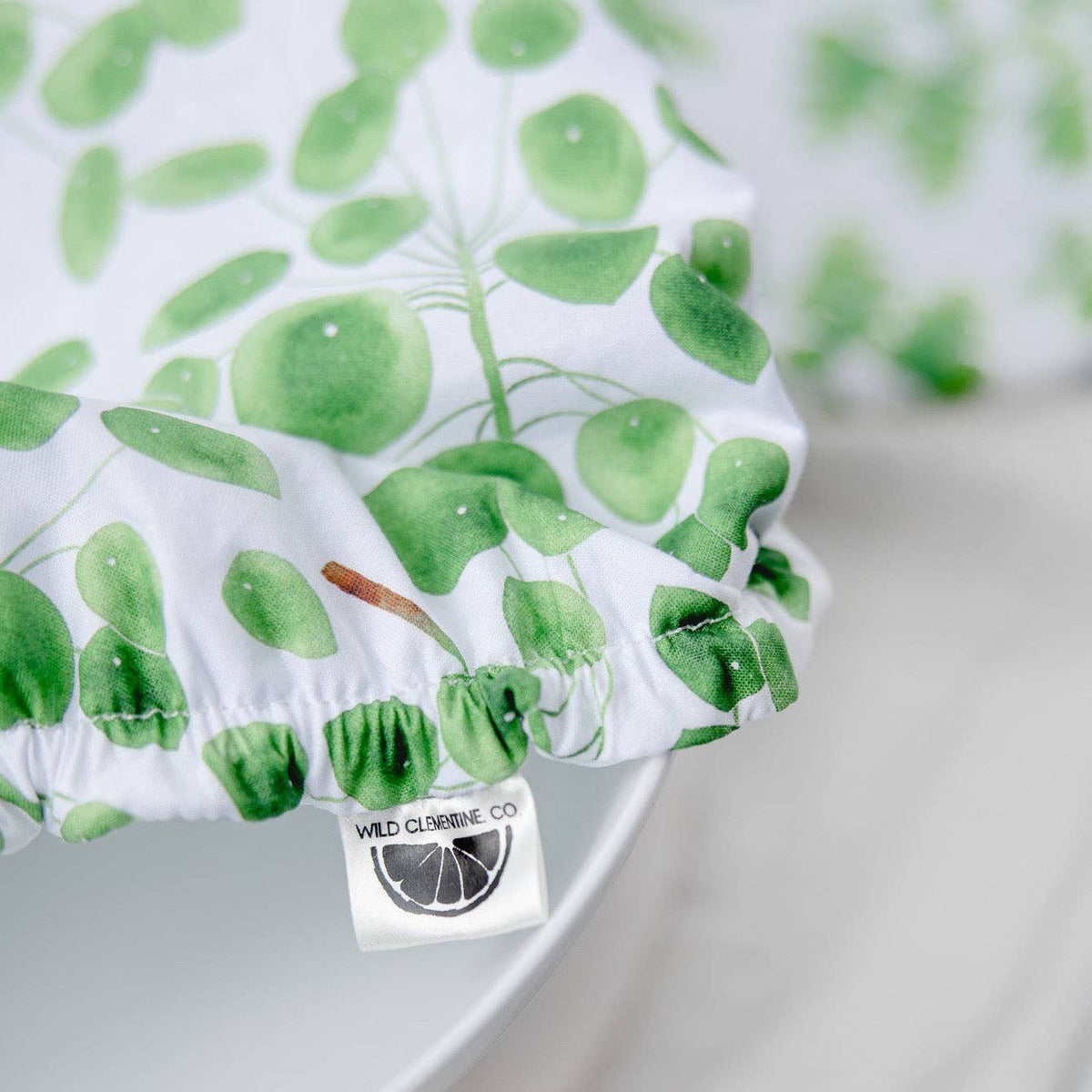 A close-up of a white fabric product with a green leaf pattern, showing a ruffled edge and a sewn-on label that reads 'WILD CLEMENTINE CO'.