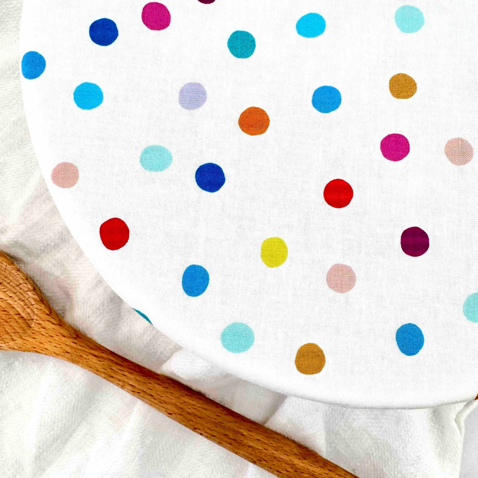 A close-up of a white fabric bowl cover, decorated with colorful polka dots in red, blue, pink, yellow, orange, and purple. A wooden spoon rests on the white fabric background.