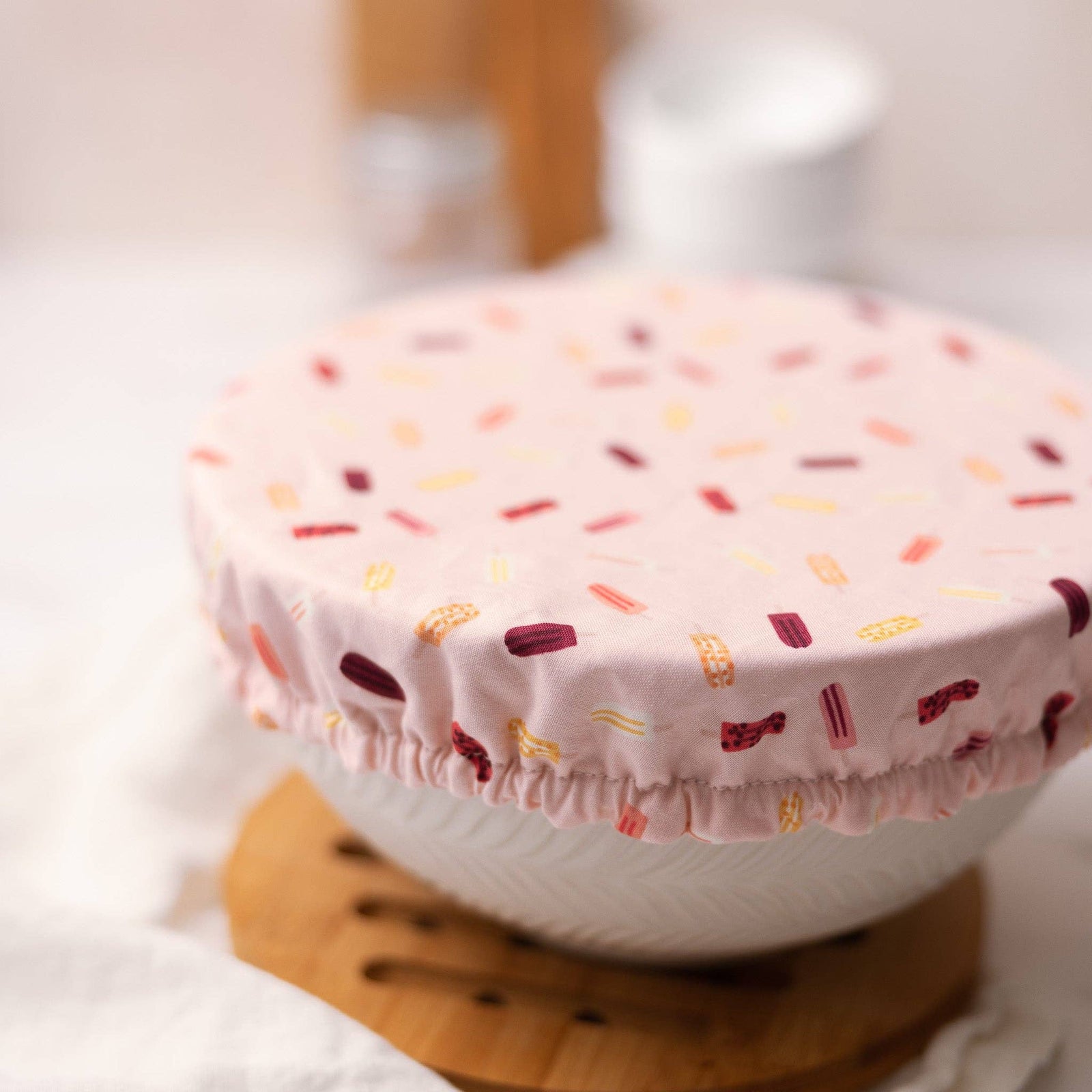 A light pink fabric bowl cover with a colorful popsicle pattern, draped over a white ceramic bowl on a wooden trivet.