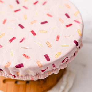 A close-up of a round fabric cover with a ruffled edge, printed with colorful popsicle motifs on a pale pink background, draped over a round wooden stand.