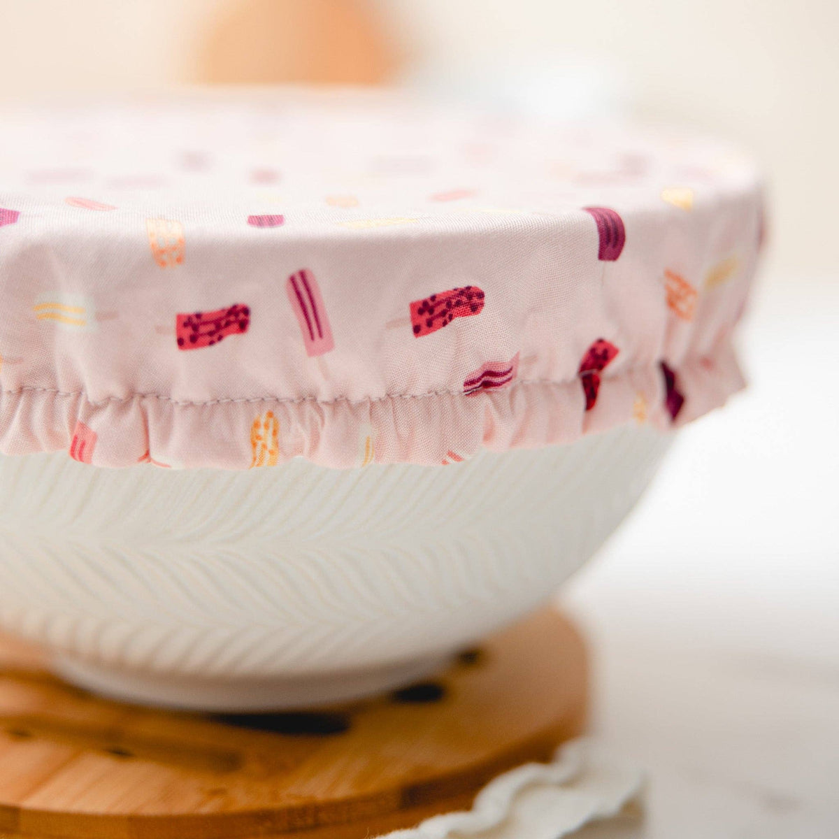 A close-up of a white ceramic bowl covered with a light pink fabric lid featuring a colorful popsicle pattern, resting on a wooden stand.