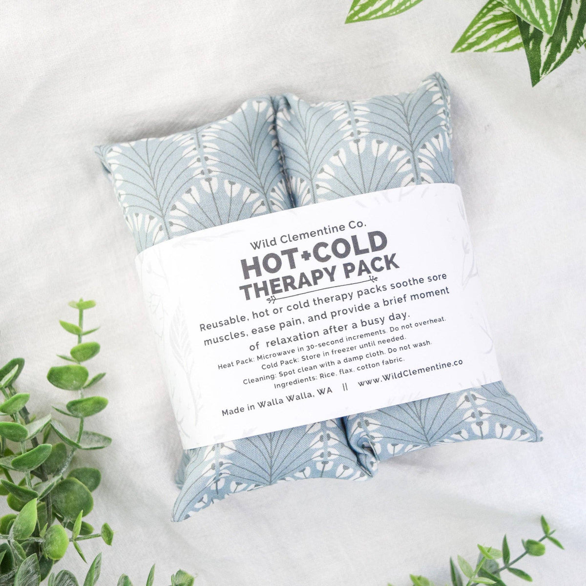 Wild Clementine Co. Hot and Cold Therapy Pack in a soft fabric pouch with a blue and white leaf pattern, wrapped in a white paper band with product details, styled on a white textured surface with green leaves.