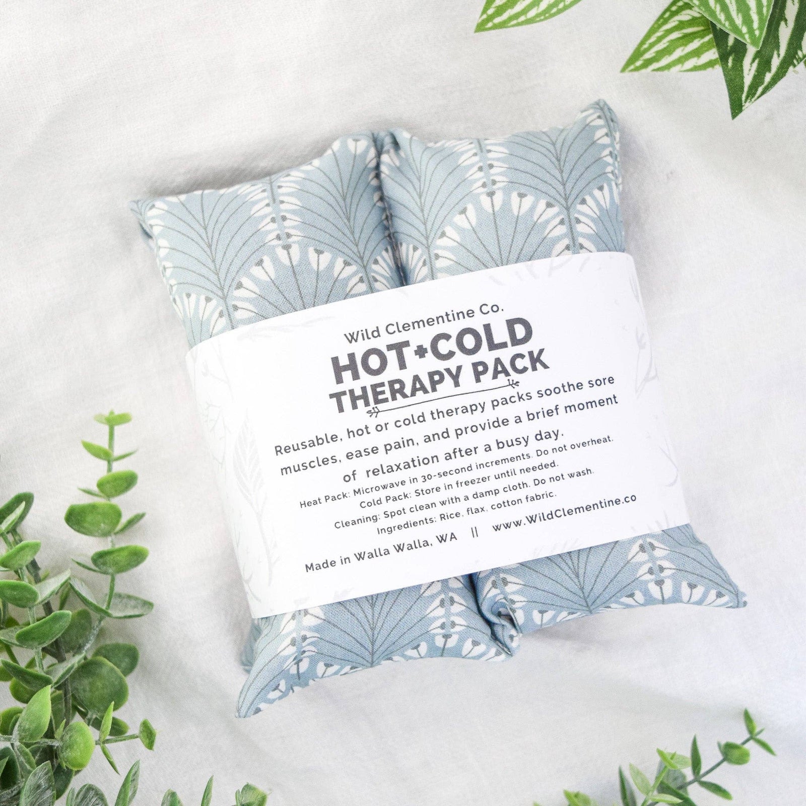 Wild Clementine Co. Hot and Cold Therapy Pack in a soft fabric pouch with a blue and white leaf pattern, wrapped in a white paper band with product details, styled on a white textured surface with green leaves.