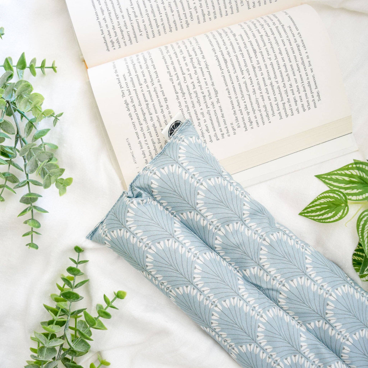 A light blue and white patterned heat pack lying on a white fabric surface next to an open book, with green leaves arranged around it.
