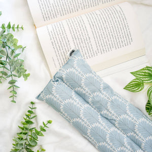 A light blue and white patterned heat pack lying on a white fabric surface next to an open book, with green leaves arranged around it.