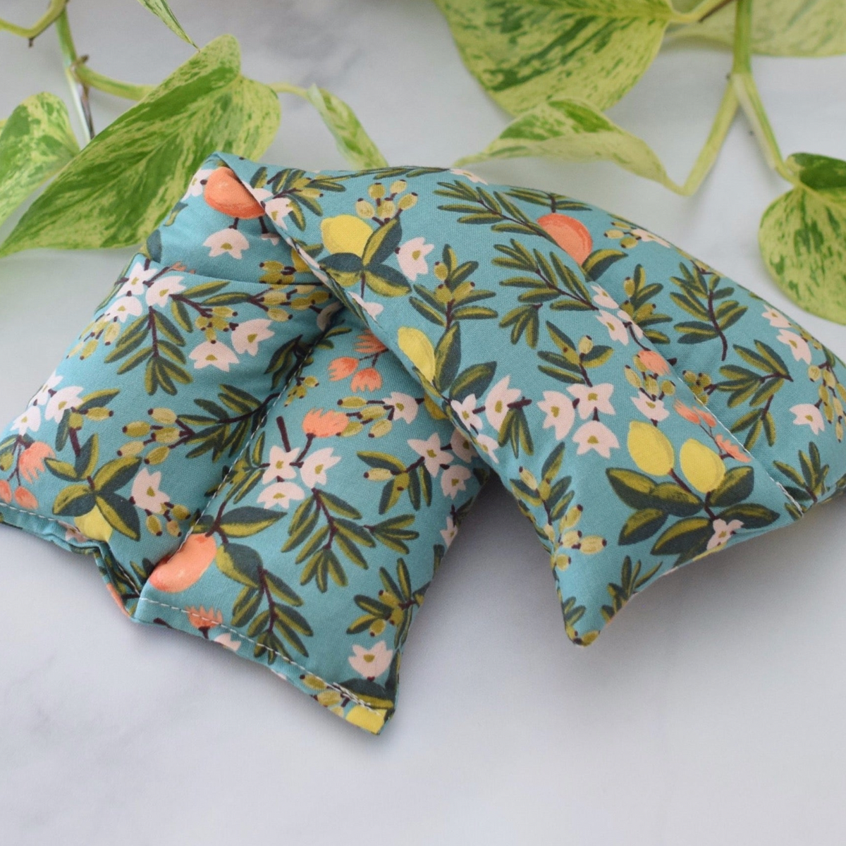 A hot/cold therapy pack with a floral print on a light blue background, styled with green variegated leaves on a white marble surface.
