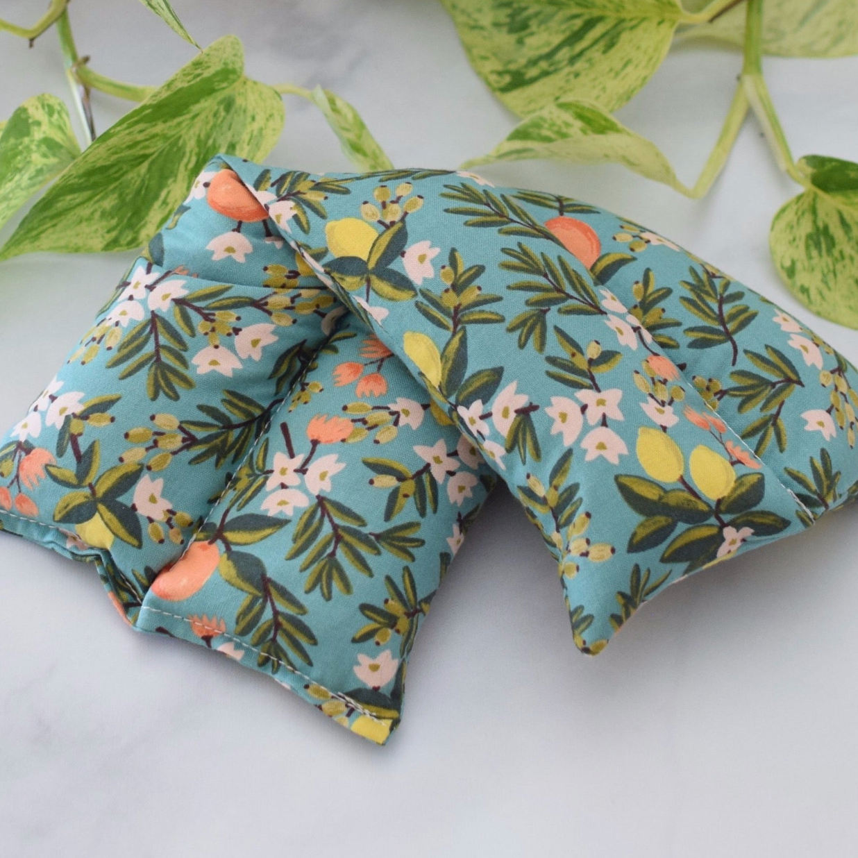 A hot/cold therapy pack with a floral print on a light blue background, styled with green variegated leaves on a white marble surface.