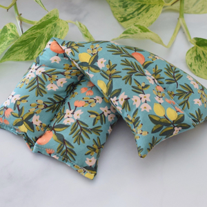 A hot/cold therapy pack with a floral print on a light blue background, styled with green variegated leaves on a white marble surface.