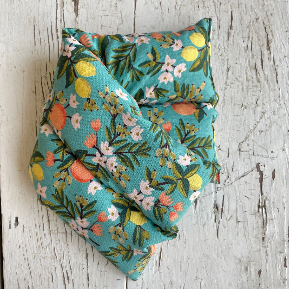 A folded hot/cold pack, featuring a vibrant teal blue-green background with a floral and citrus print of lemons, oranges, and white blossoms. It is displayed on a rustic, whitewashed wooden surface.