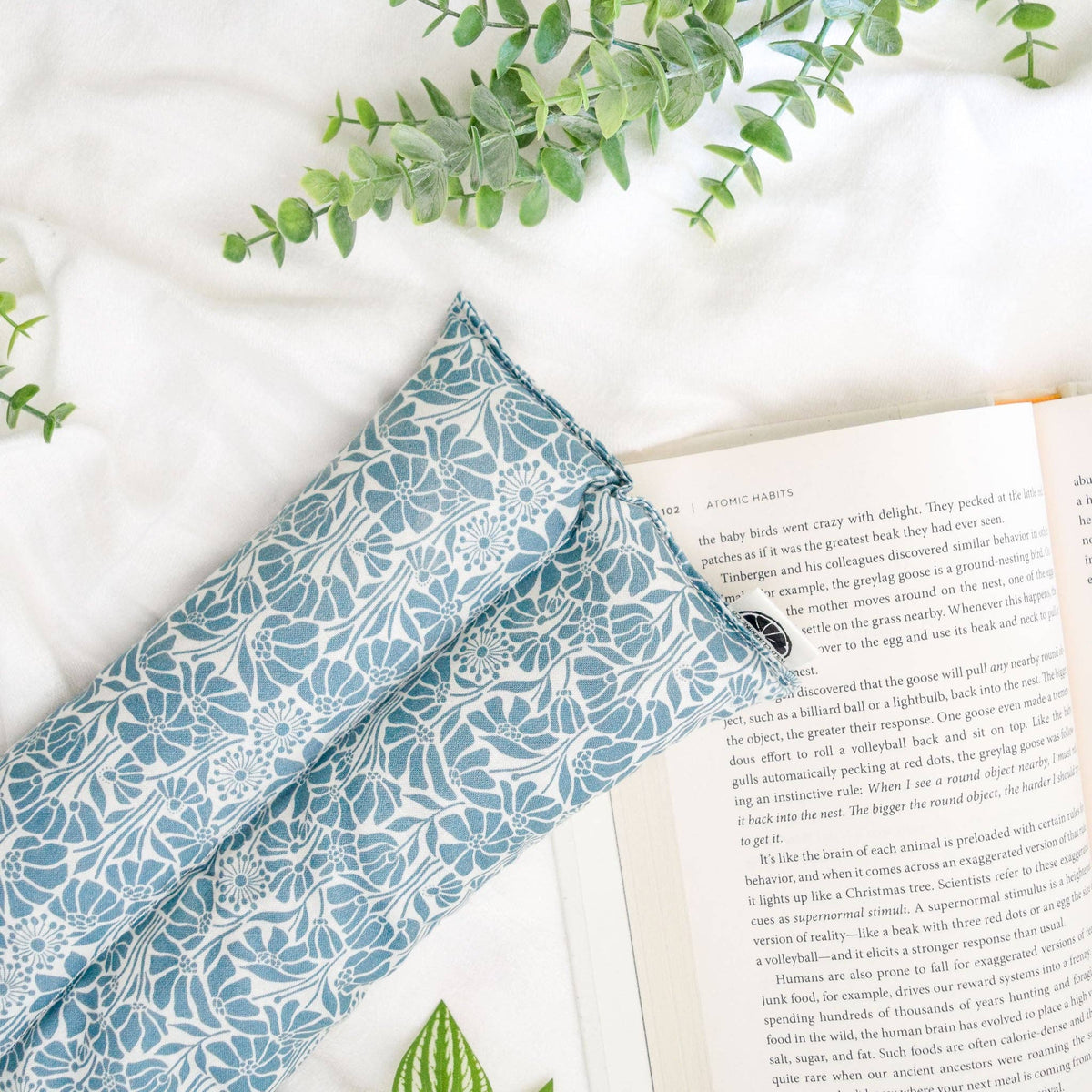 Blue floral hot/cold pack on a white surface with an open book and green leaves.