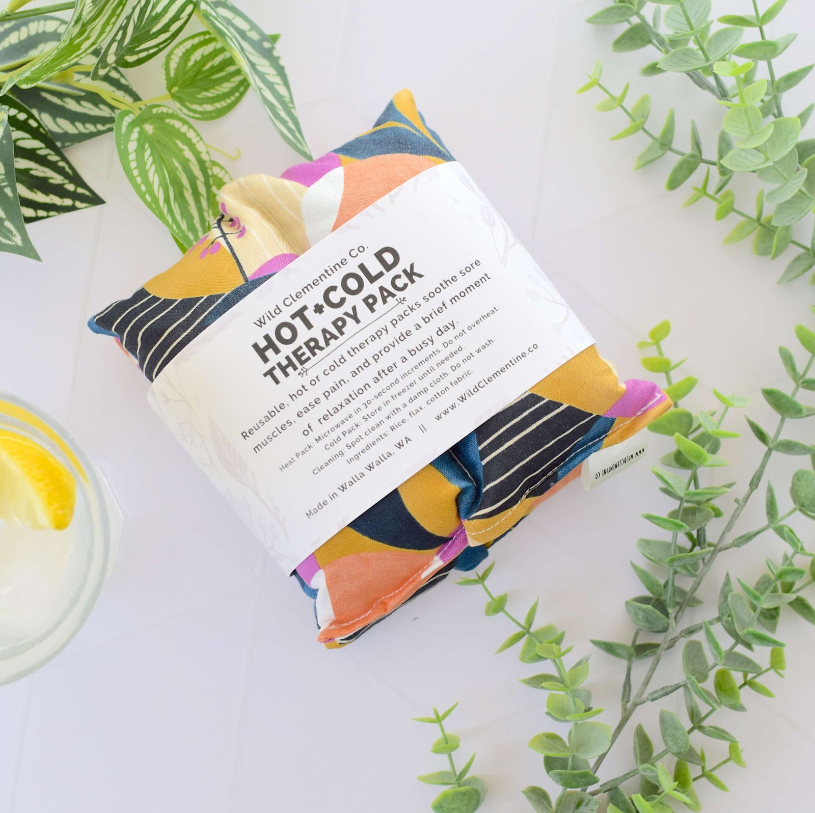 A folded hot-cold therapy pack from Wild Chamomile Co. with a white paper label, styled on a white surface with green leaves and a glass of lemon tea.