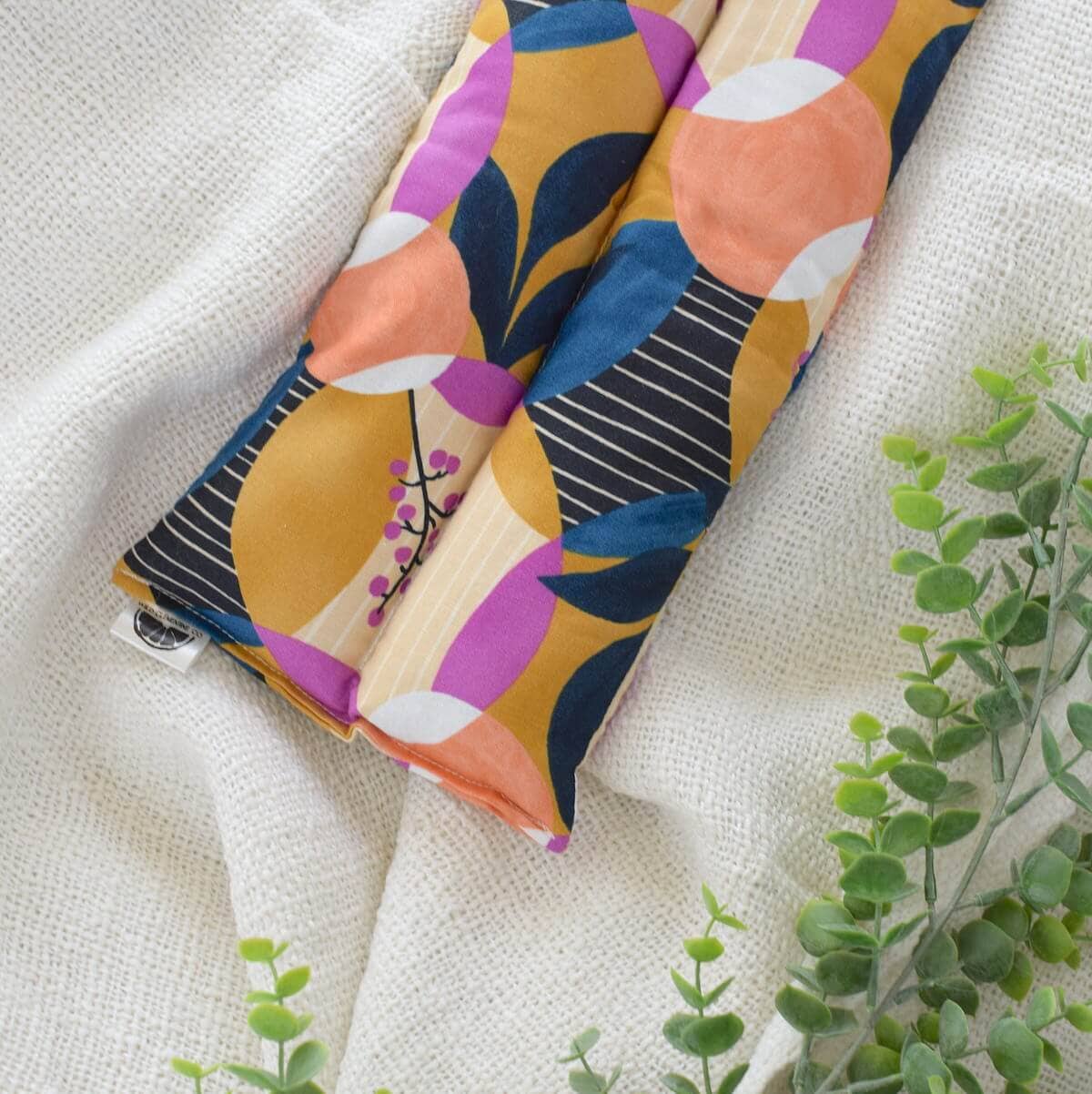 A folded hot/cold pack displayed on a textured cream fabric background with green plant stems around it. The product features a bold abstract print with mustard yellow, navy blue, purple, peach, white, and black elements.