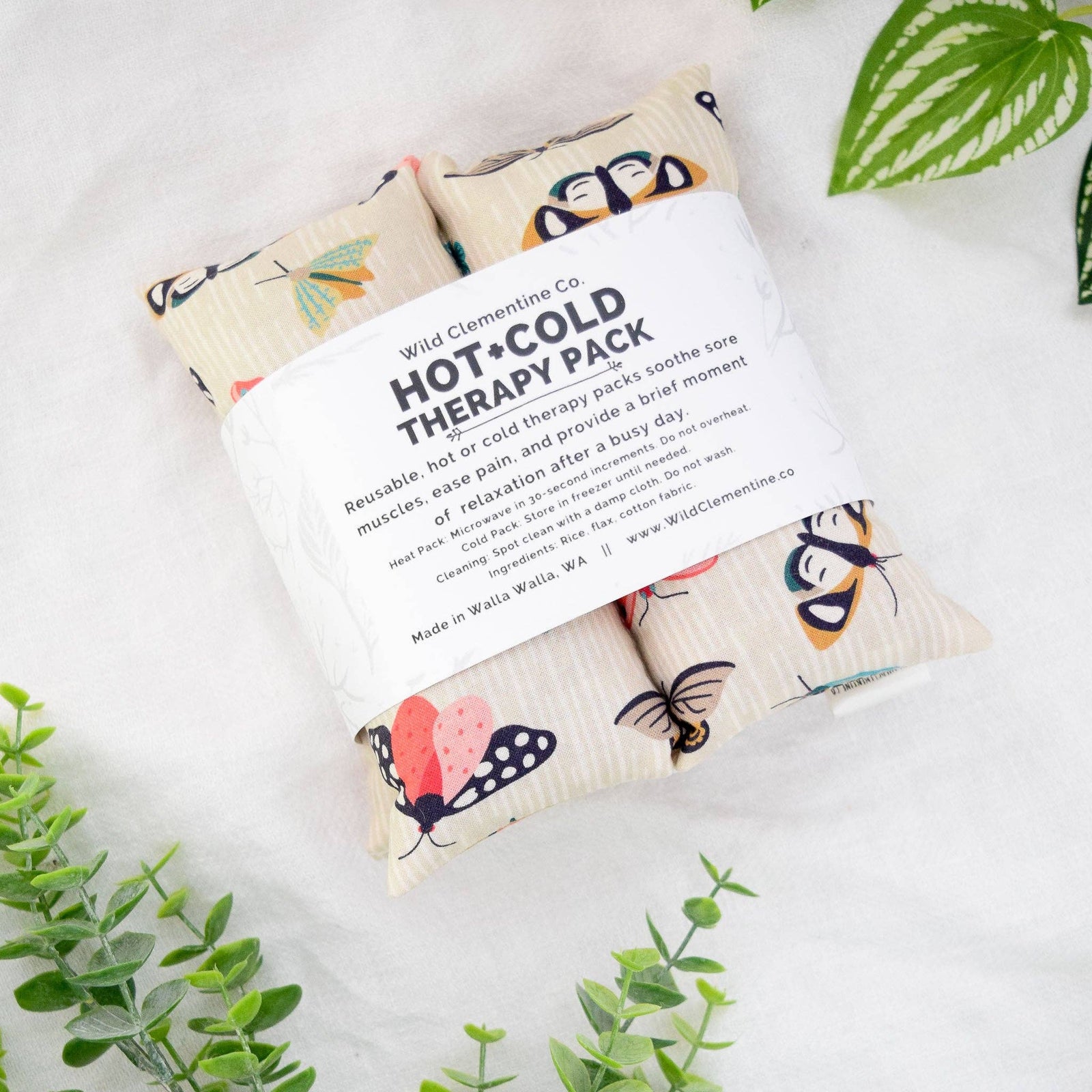 A hot and cold therapy pack by Wild Clementine Co, featuring three fabric pouches with colorful moth illustrations, wrapped in a white paper band, styled on a white textured fabric with green leaves.