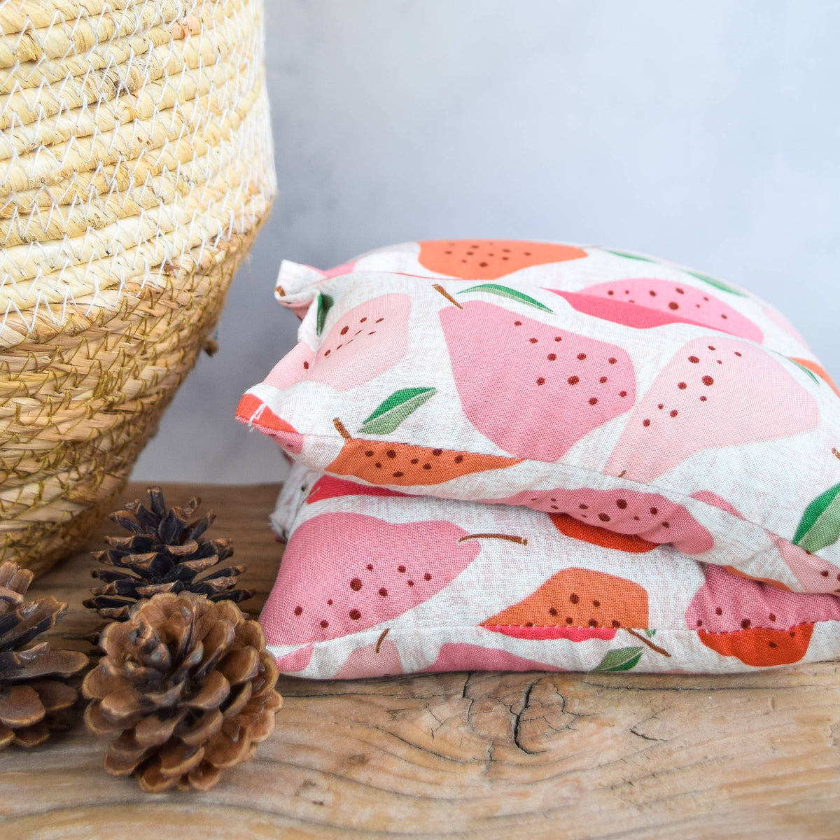 A folded hot/cold pack with a pink and orange pear pattern on a white background, styled on a wooden surface next to pinecones and a woven basket.