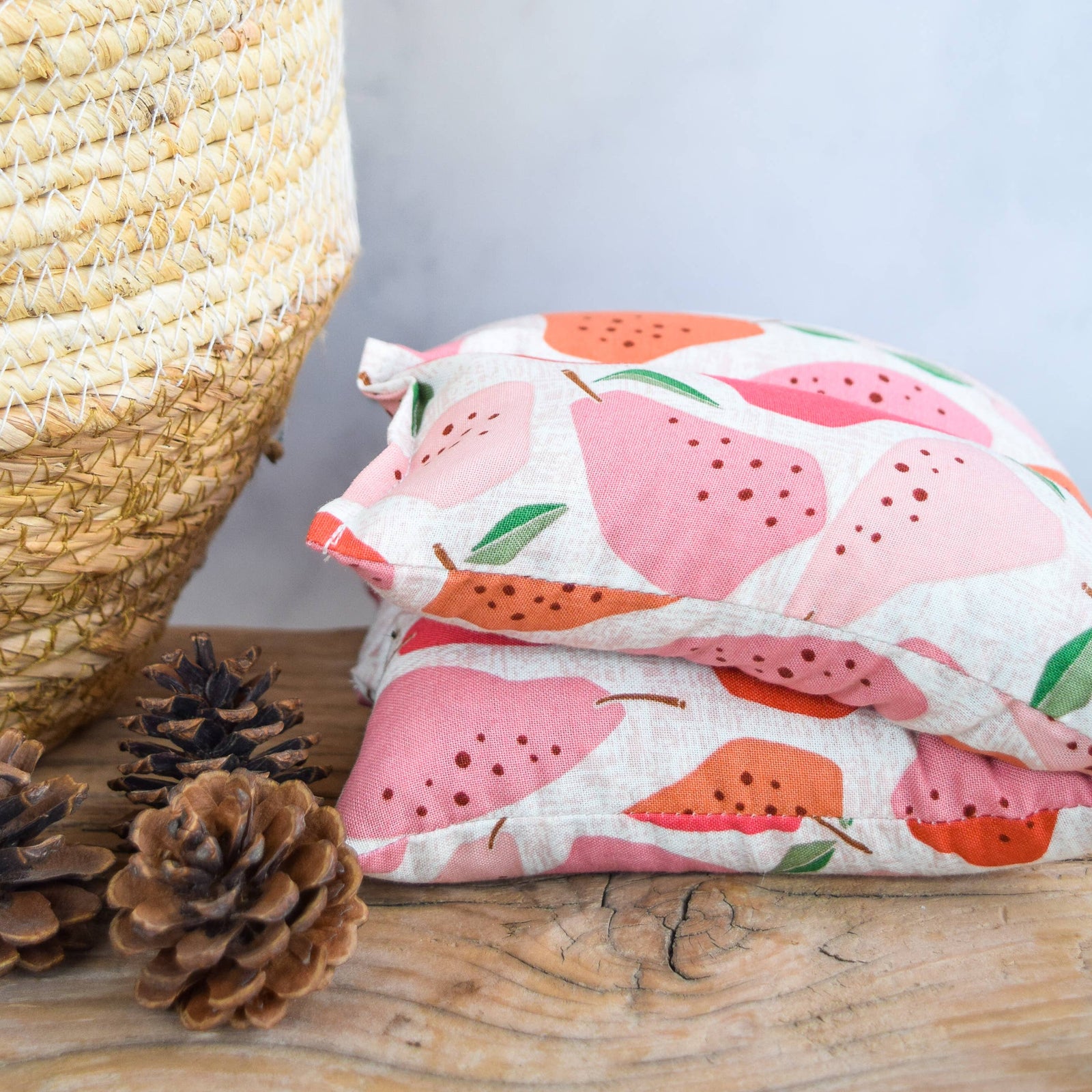 A folded hot/cold pack with a pink and orange pear pattern on a white background, styled on a wooden surface next to pinecones and a woven basket.