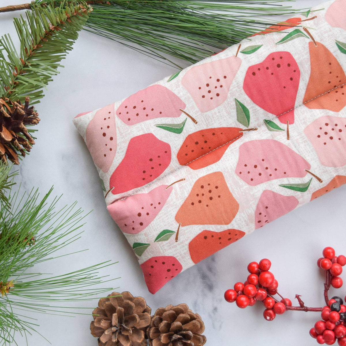 A hot/cold pack with a pear pattern in shades of pink, coral, and orange on a cream linen background, styled on a white marble surface with pine branches, pinecones, and red berries.