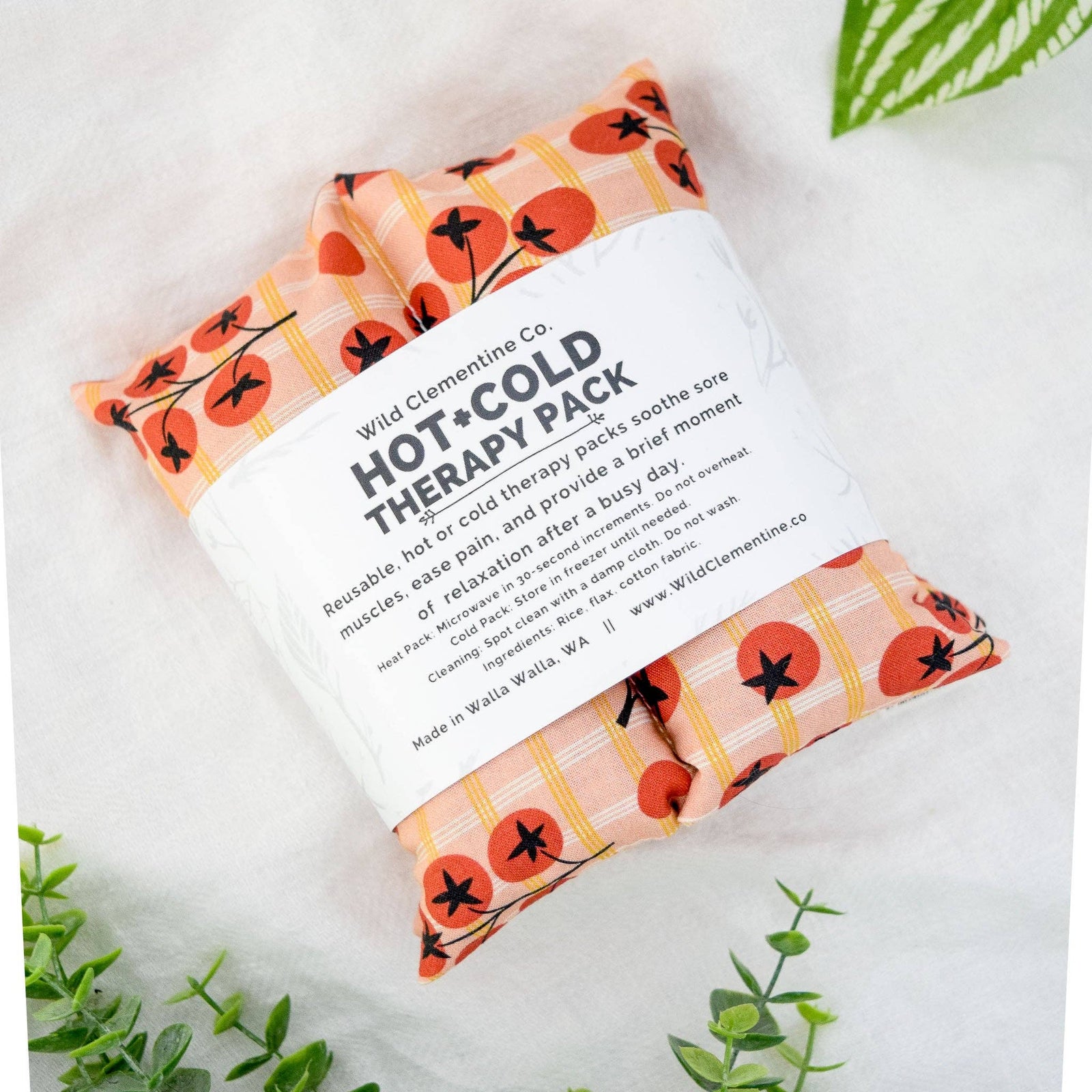 Wild Clementine Co. Hot + Cold Therapy Pack in a reusable cotton pad with a white paper band label, styled on a white textured surface with green leaves.