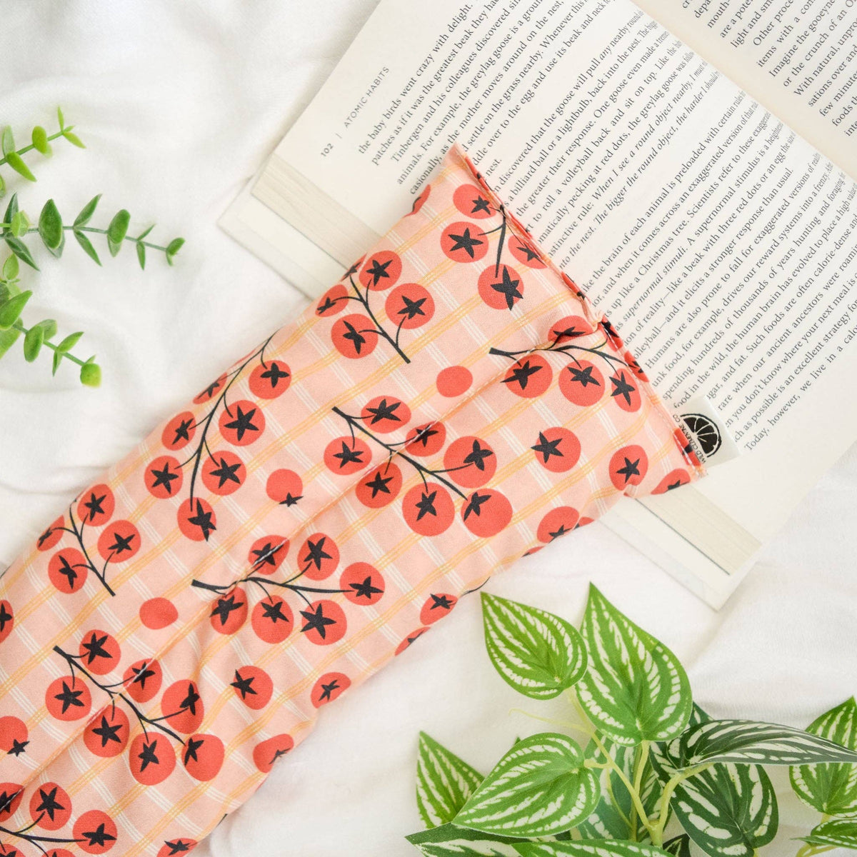 A hot/cold pack with a peach and orange plaid pattern and red tomato motifs, styled on a white fabric surface next to an open book and green plants.