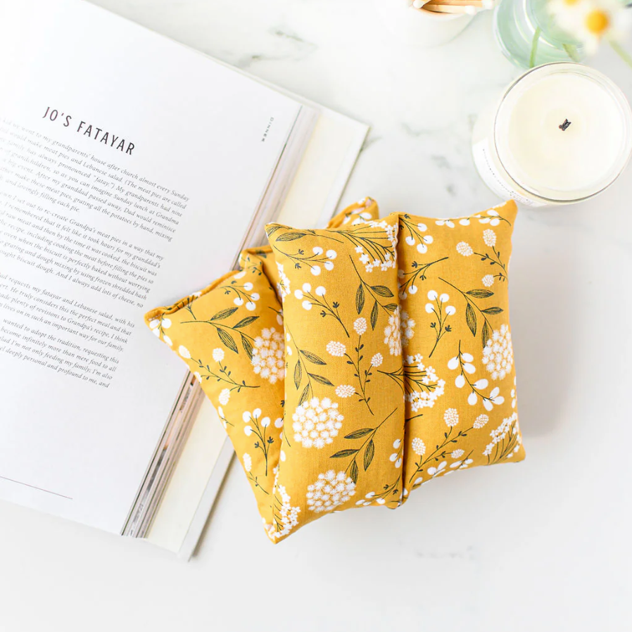 yellow floral-patterned hot/cold pack on a white surface with an open book and candle in the background.