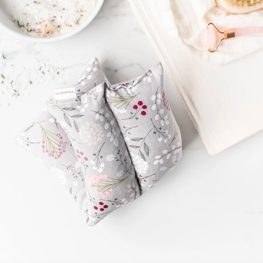 Floral-patterned hot/cold pack on a white surface with a bowl of bath salts and a book in the background.