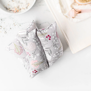 Floral-patterned hot/cold pack on a white surface with a bowl of bath salts and a book in the background.