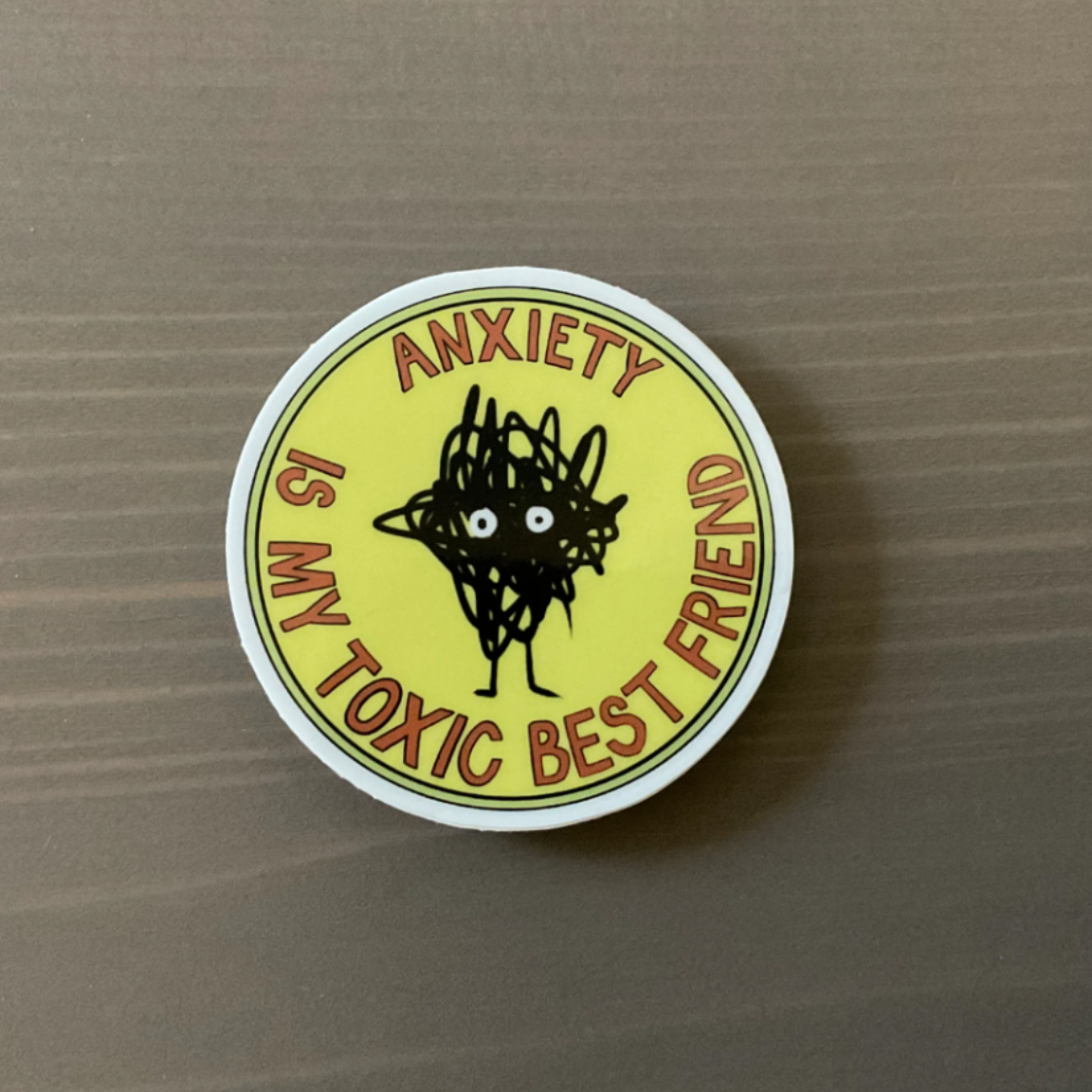 A round sticker that has a scribble with eyes in the center and says "Anxiety is my toxic best friend"